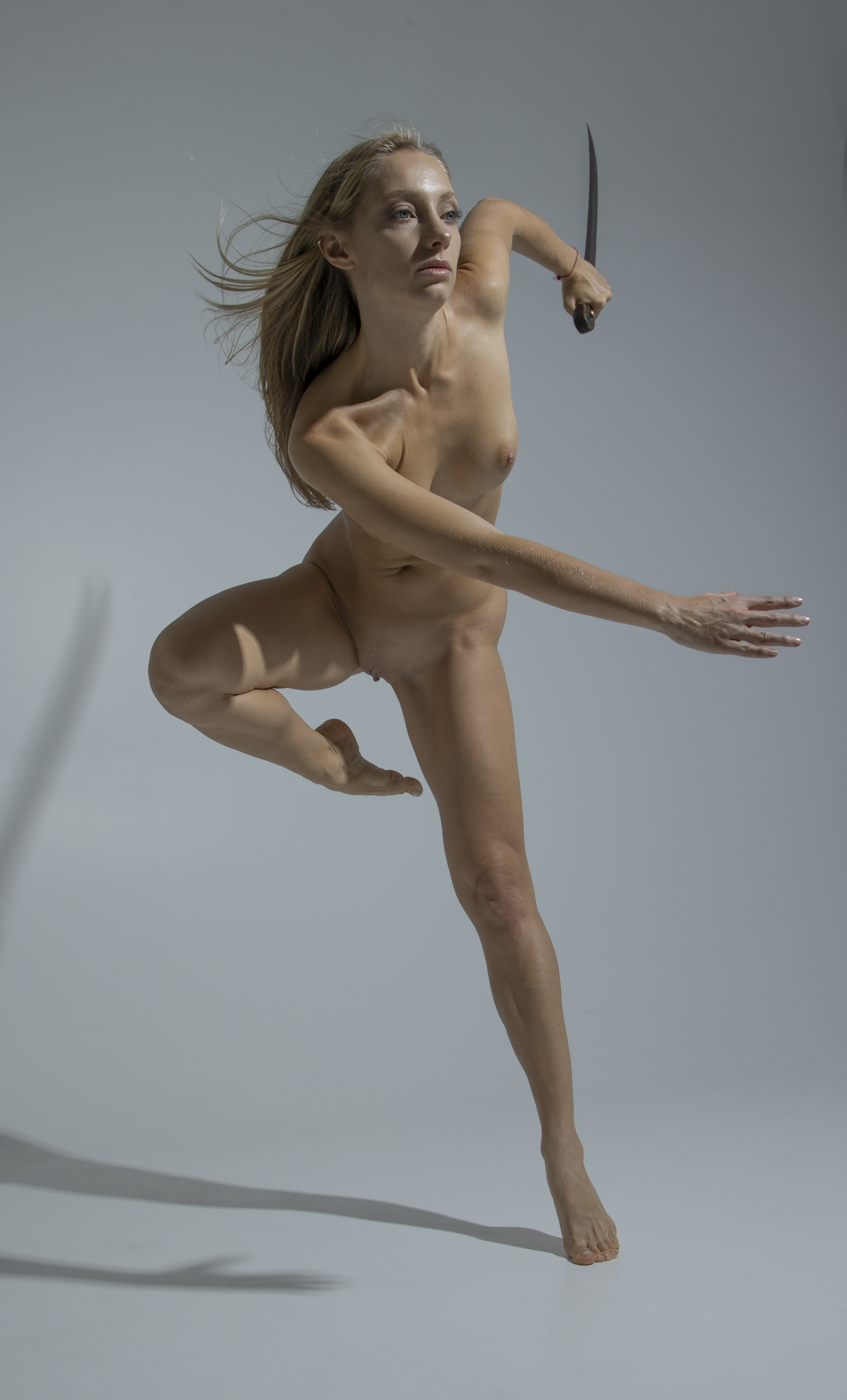a nude woman with blonde hair is captured in a dynamic studio shot. she is holding a knife in one hand while dancing or jumping. the image is full length, showcasing her entire body in motion against a neutral backdrop.