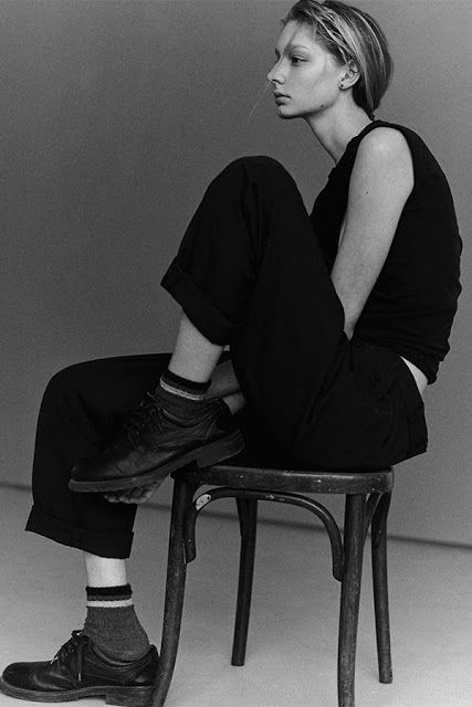 a young woman in dark clothing and socks is sitting on a wooden chair with her legs crossed. she is leaning forward slightly and looking off to the side with a serious expression. the photo is in black and white.