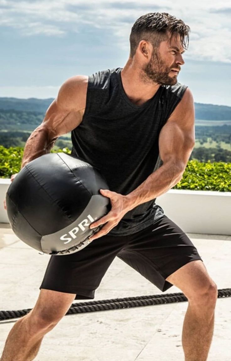 a muscular man in a black tank top and shorts is holding a medicine ball during a workout outdoors. he is on a patio with grass in the background.
