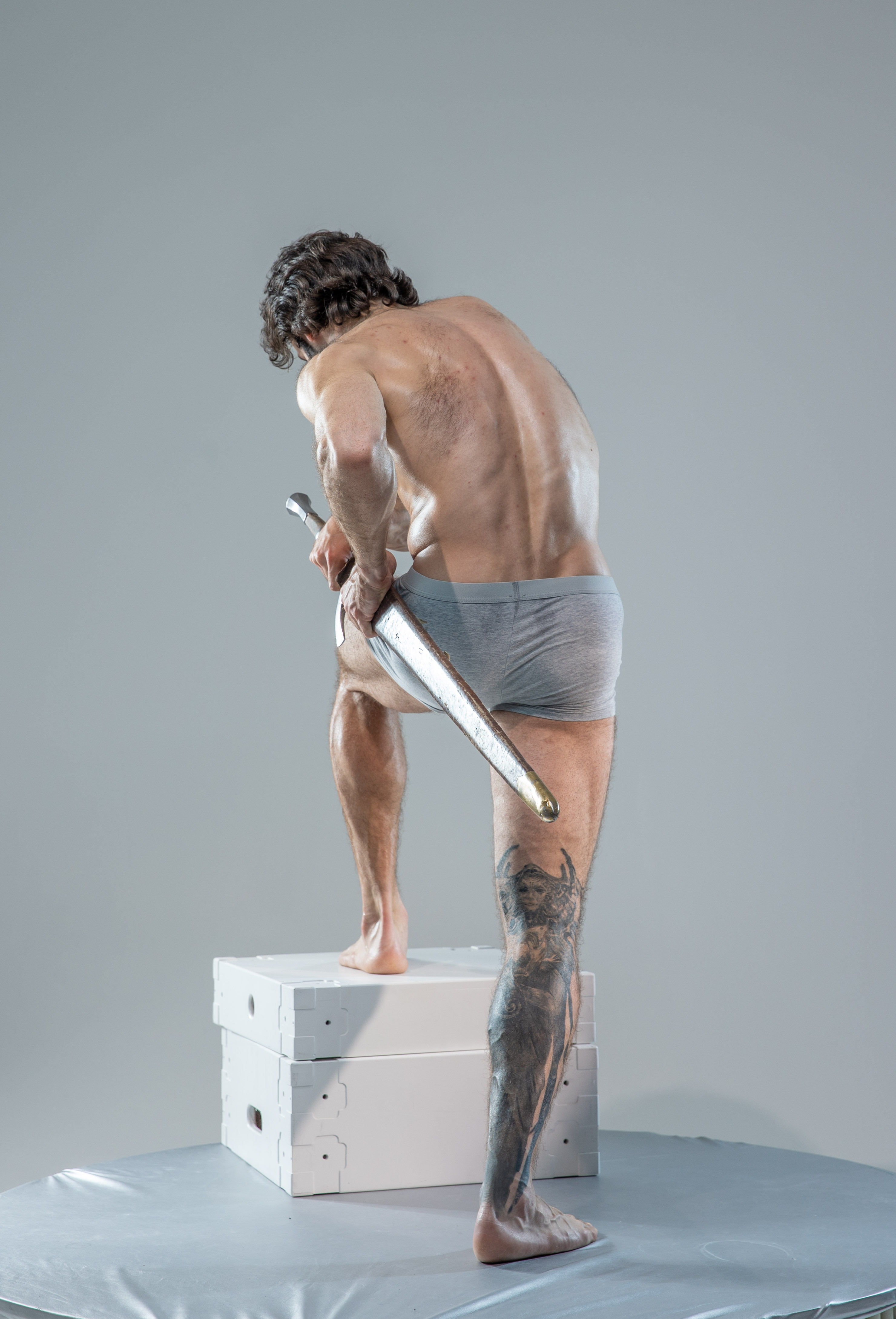 a rearview studio shot of a muscular man with a dragon tattoo, holding a sword in a warrior pose. the model is illuminated with soft lighting, highlighting his physique against a neutral gray backdrop. he is standing on a white platform, suggesting a controlled environment for artistic reference.