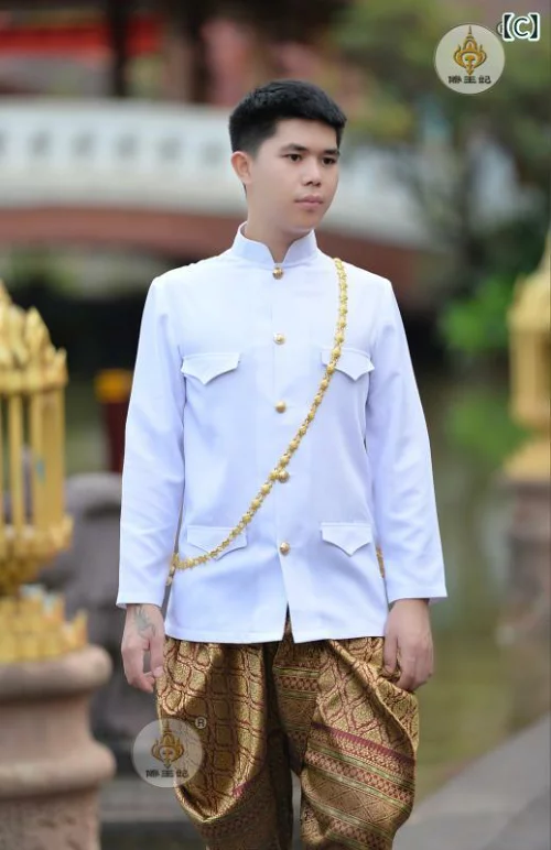 a young man in a traditional white thai outfit, with a gold chain and patterned pants, stands outside with a serious expression.