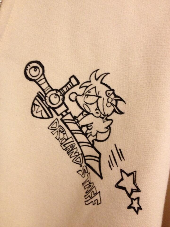 the image shows a beige jacket with a blackandwhite cartoon character and "dreamland staff" text printed on it. the cartoon features a spikyhaired character with horns and a bandage under their eye clinging to a large stylized sword. there is also a simplified fivepointed star.