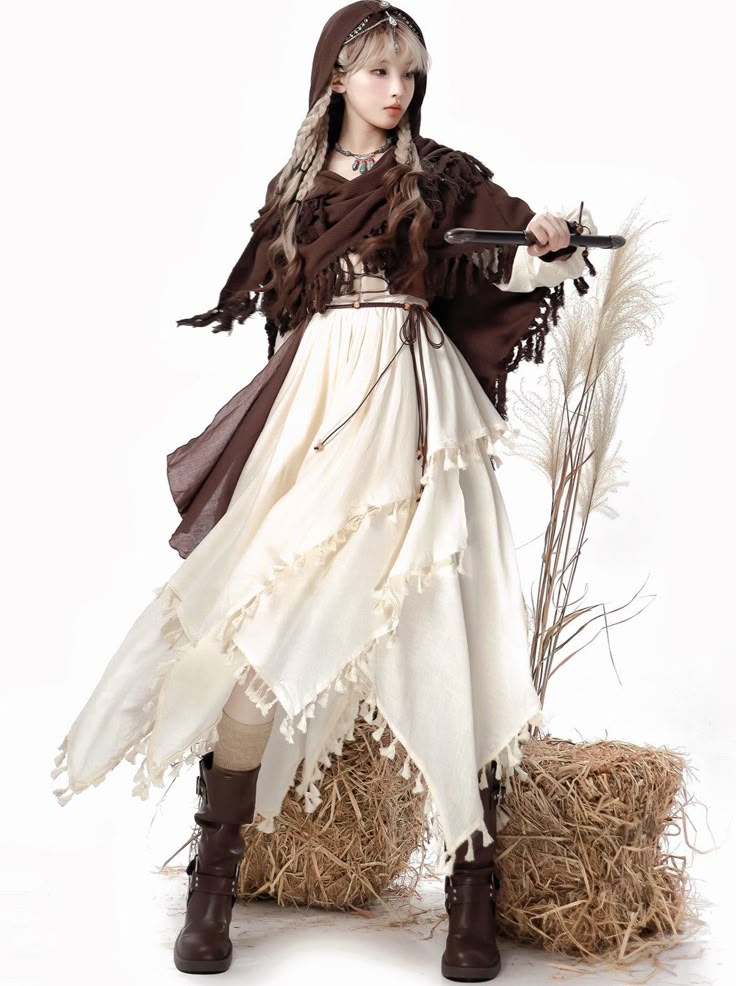 a woman is wearing a brown hooded shawl and a white dress with tassels. she is holding a dark object in her right hand. she is also wearing brown boots. there are two straw bales and some dried grass around her. the background is white.