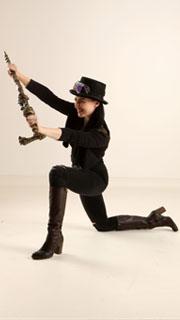 a woman in a steampunkinspired outfit strikes an action pose. she is holding a unique weapon and wears a hat with goggles, a black jacket, tight pants, and brown boots. the setting appears to be a studio with a plain white background and soft lighting.