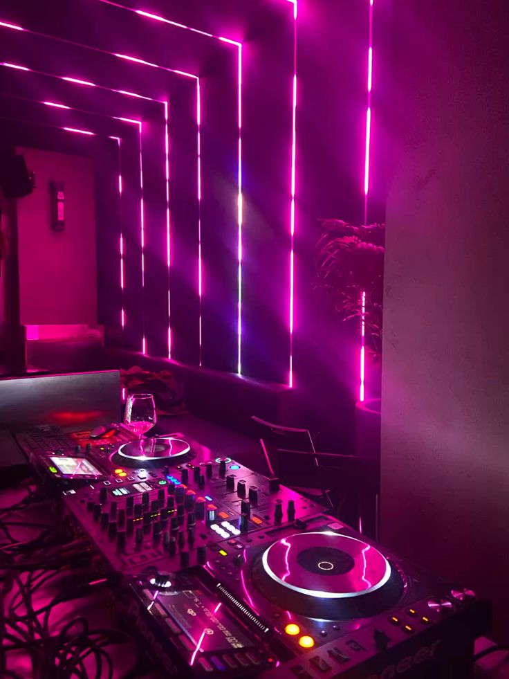 the image showcases a professional dj setup in a nightclub setting. two dj turntables are prominently displayed, wired to a mixer with various knobs and buttons, all illuminated by vibrant pink and purple neon lights that create a futuristic and energetic ambiance. the background features a hallwaylike structure with the same neon lights, emphasizing depth and leading the eye towards the back of the room. a single wine glass sits atop the equipment, hinting at a momentary pause in the otherwise lively atmosphere.