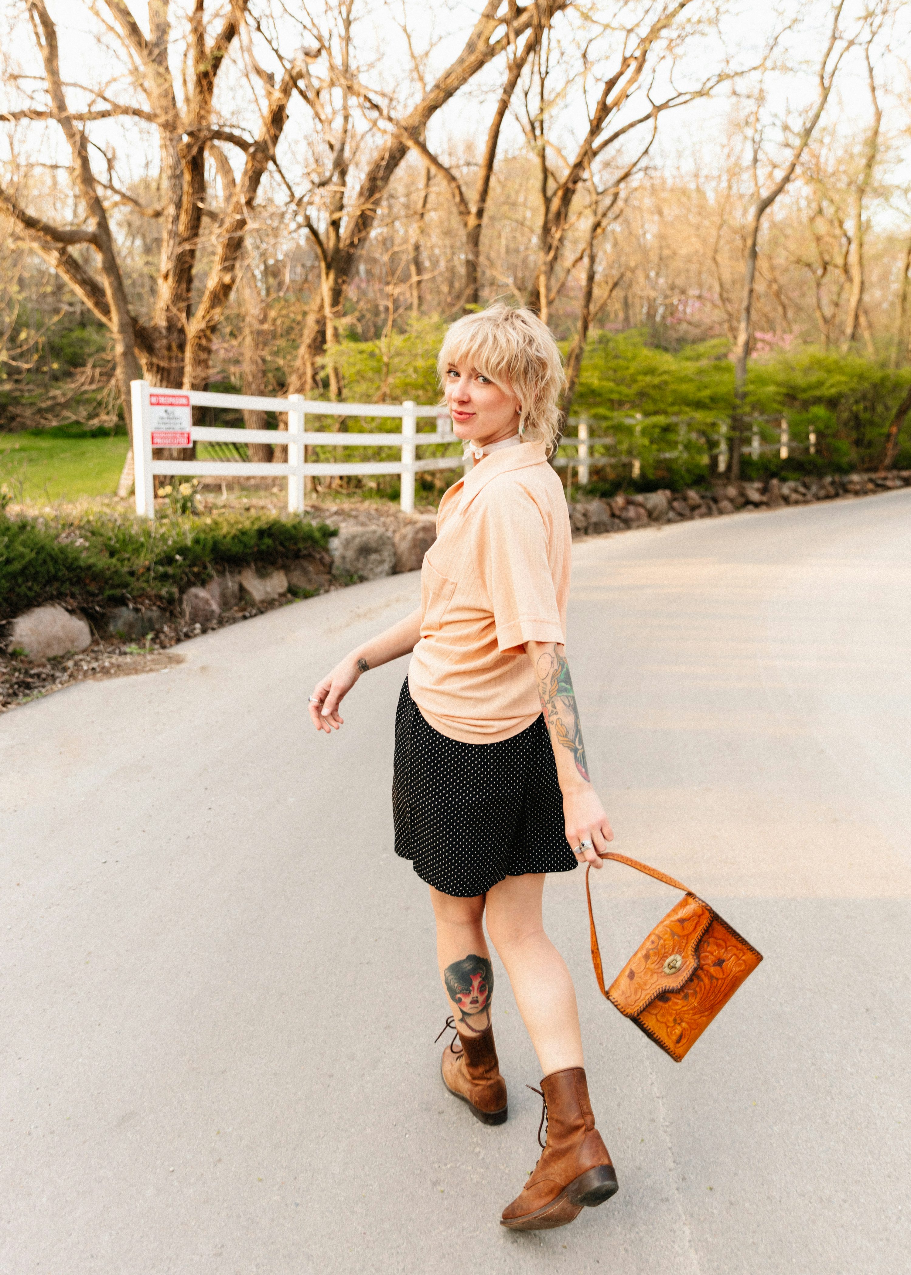 a young woman with short blonde hair and tattoos walks down a country road. she is wearing a beige shirt, a black polka dot skirt, and brown boots. she carries a vintage leather handbag. the setting is outdoors, with trees and a white fence in the background. the overall style is casual and retro.