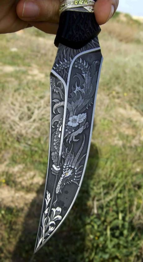 a hand holds a highly ornate and engraved knife or dagger. the blade is silver and features a detailed dragon design intertwined with floral patterns. the hilt of the knife appears to be decorated with a ring and potentially some gemstone accents. the background is slightly blurred, suggesting an outdoor setting with green vegetation.