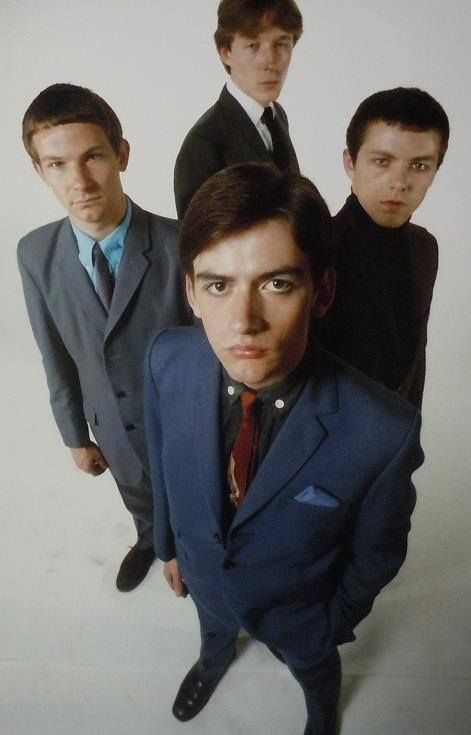 a group of four men are standing in suits, looking at the camera. they look like a band from the 1970s. two of the men are wearing blue suits, one is wearing a brown suit, and the other is wearing a black suit.