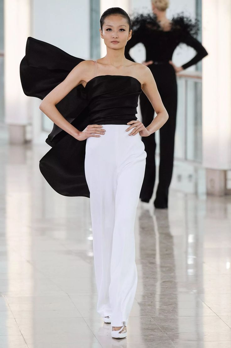 a fashion model walks the runway in a white pantsuit and a black top with a bow. she is wearing high heels and has her hair pulled back. the model is posing for the camera and looking confident. the background is blurred and out of focus.