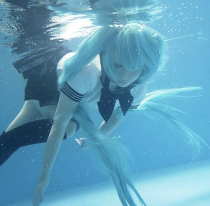 an underwater image of a woman dressed in a cosplay outfit, specifically a school uniform or sailor outfit. she has long, flowing blue hair (possibly a wig) and is submerged in clear blue water, likely in a swimming pool. the uniform consists of a white top with a black bow and a dark skirt. the overall image has a dreamlike, ethereal quality.