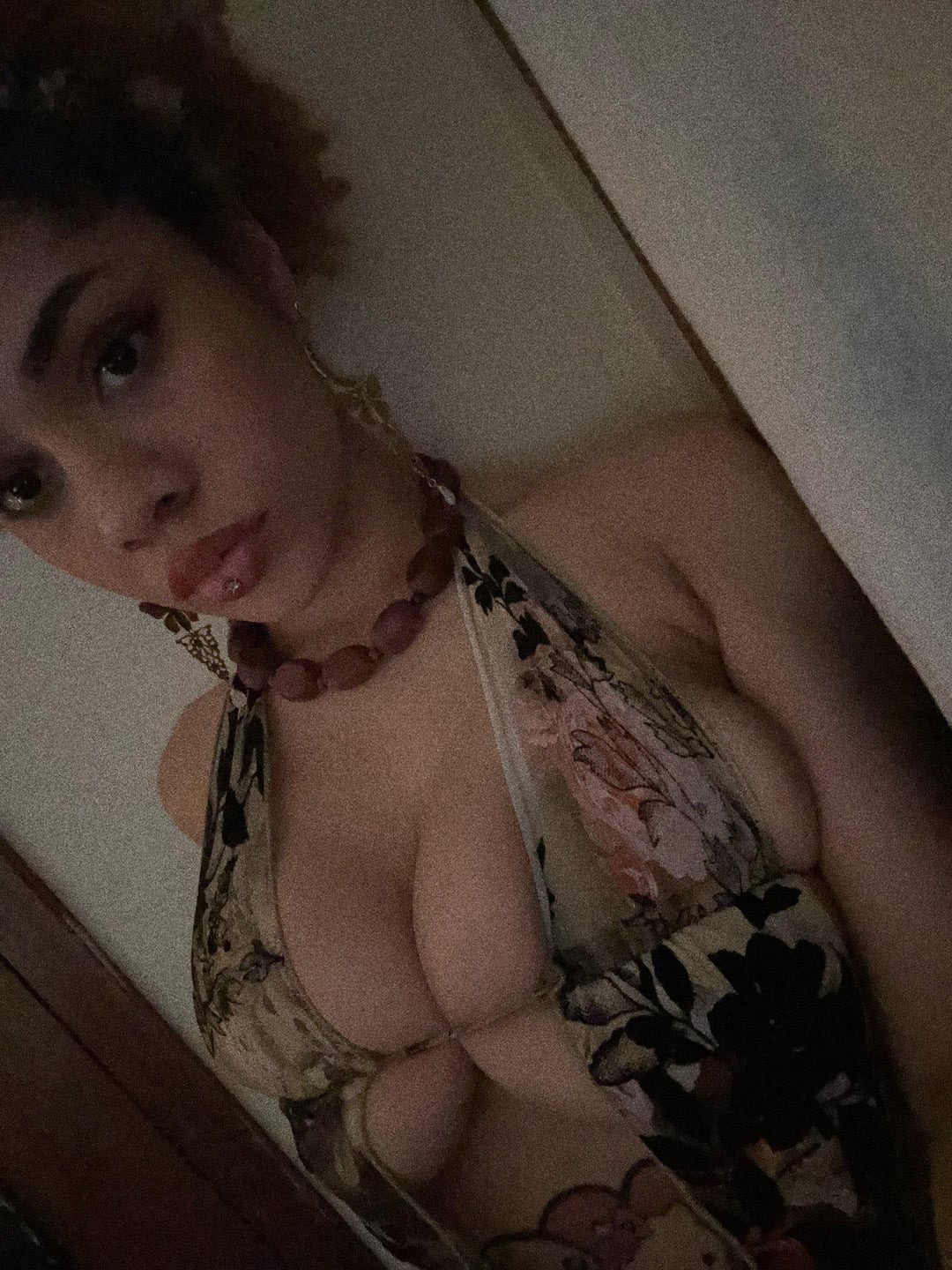 a woman with dark hair in a halter top with a floral print and bead necklace is looking at the camera. she has a pierced lip and a tattoo can be seen on her chest.