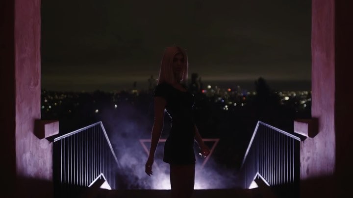 a woman in a black dress is standing at the bottom of a set of stairs outside at night. the stairs have a metal railing. the city lights are visible in the background. there is a hazy or smoky effect around the bottom of the stairs. the scene has a dark and mysterious atmosphere. the architectural elements suggest a building or structure, possibly made of concrete.