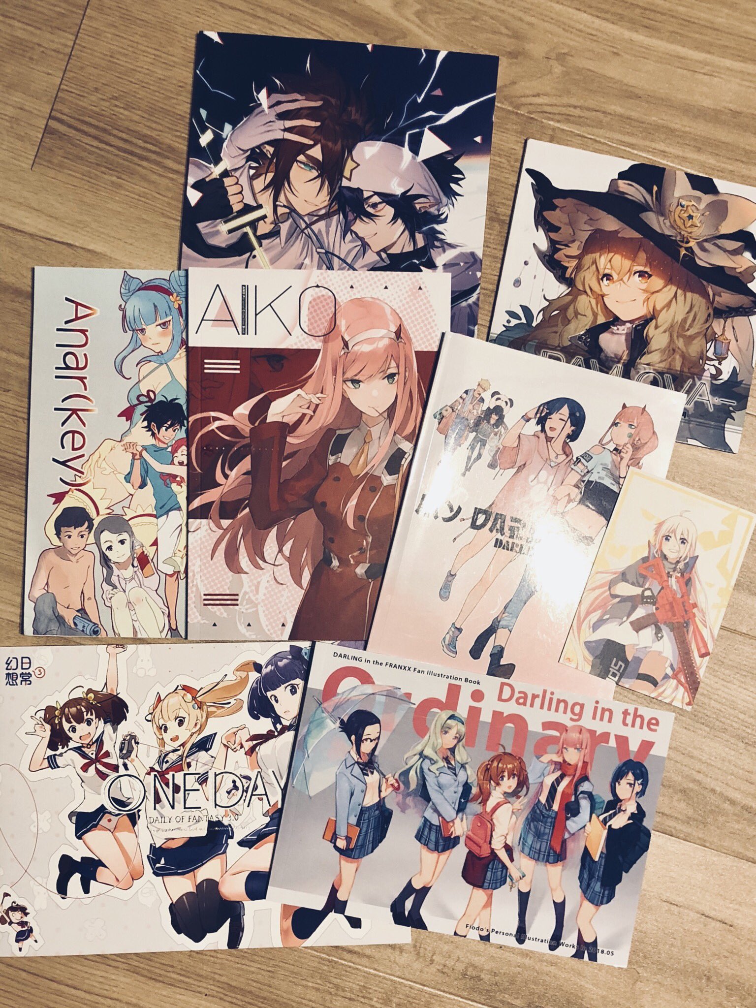 the image shows a collection of seven anime art books splayed out on a light brown wooden floor. the books feature colorful covers with various anime characters in different poses and outfits. the art style is distinctly japanese manga and anime, with large eyes, vibrant hair colors, and dynamic compositions.  the lighting suggests an indoor setting illuminated by artificial light, giving the image a slightly warm tone.
