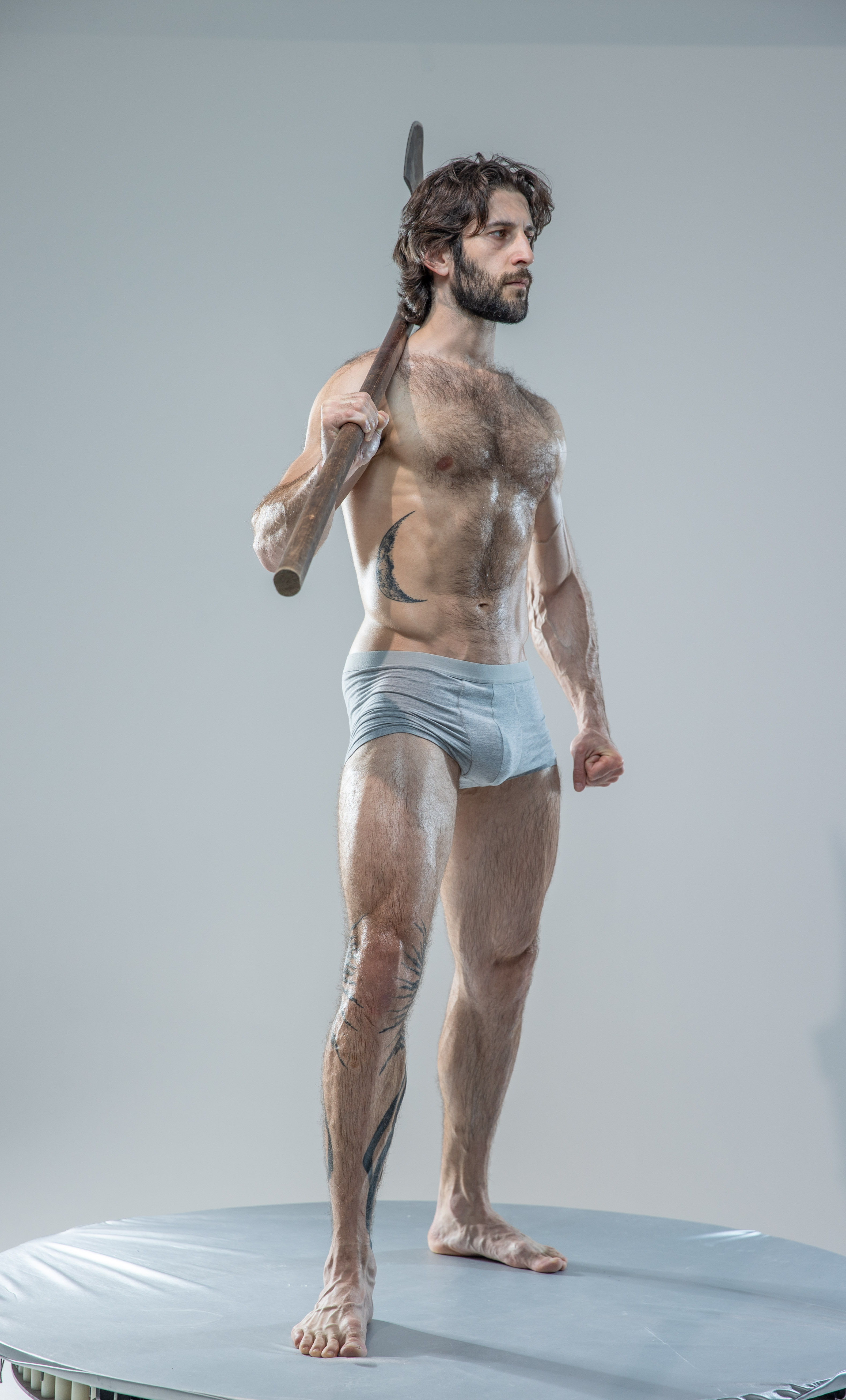 a muscular, hairy man stands on a circular platform, holding an axe over his shoulder. he wears only underwear, showcasing his physique and tattoos. the studio lighting highlights his muscles and creates a dramatic atmosphere.