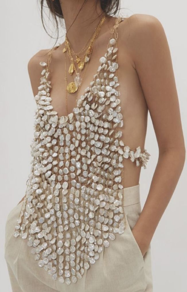 a woman is shown wearing a pearl top, several gold necklaces, and beige linen pants. she is standing and has her hands in her pockets. the focus is on the jewelry and clothing.