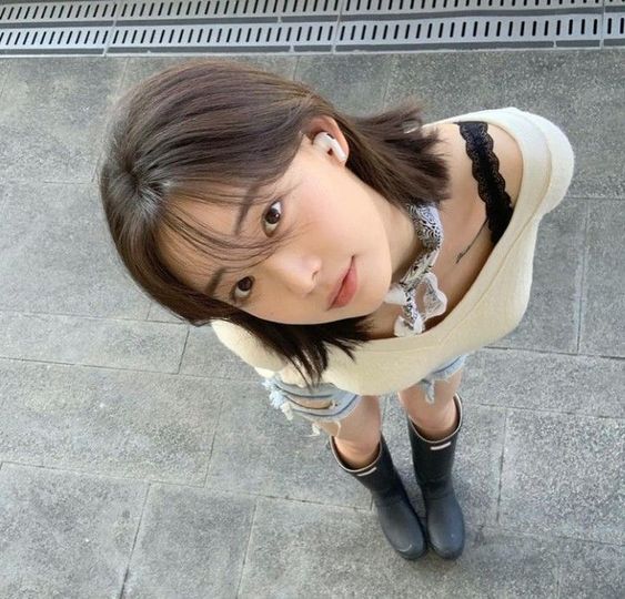 the image shows a young woman taking a selfie from a high angle, looking down at the camera. she has short dark hair, is wearing a white shirt, denim shorts, and black rain boots. she is also wearing a small patterned scarf and white wireless earbuds. the background is a gray tiled sidewalk. the photo is taken outdoors in natural daylight.