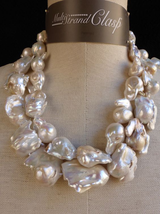 a closeup shot of a multistrand baroque pearl necklace on a mannequin. the necklace features large, irregularly shaped white pearls with a luminous sheen. the clasp is hidden at the back, emphasizing the natural beauty of the pearls. the background is a neutral beige, allowing the pearls to be the focal point of the image.