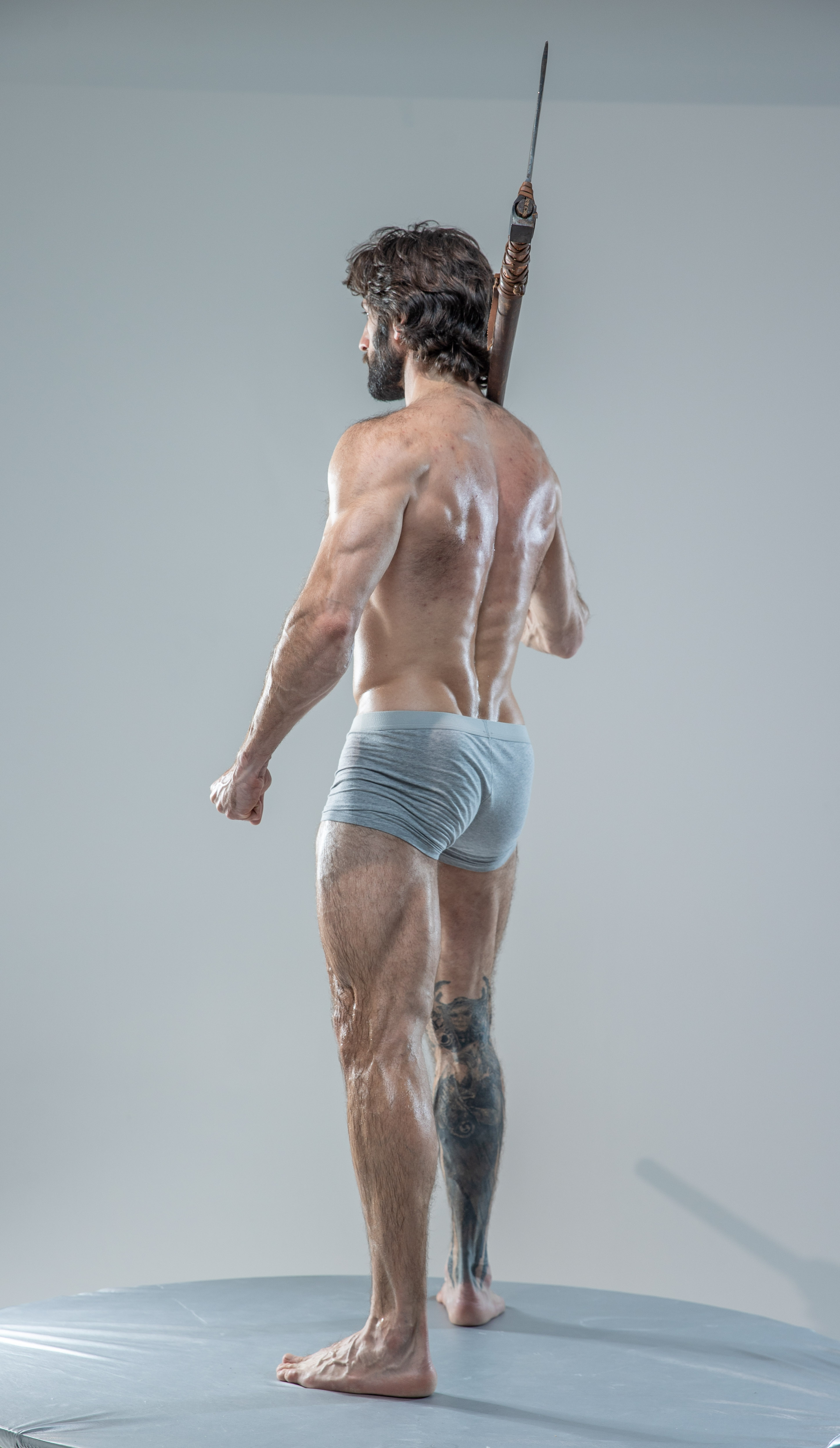 a muscular man with long hair and a beard stands on a grey platform with his back to the camera. he is shirtless, wearing only grey underwear, and has a tattoo on his left leg. he holds a spear over his right shoulder, his hand gripping the shaft. the image is a studio shot against a plain grey background.