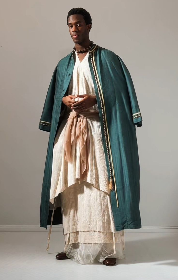 a fullshot studio portrait shows a young, darkskinned man standing and wearing a long, elaborate outfit. he faces the viewer and his hands are clasped at his waist. he wears a long, layered offwhite tuniclike garment that extends to his ankles. around his waist is tied a wide, draped tan sash. over this, he wears a long, tealcolored overcoat or robe with gold trim at the edges and cuffs. he also wears a dark beaded necklace. the background is a neutral, light gray tone, and he is wearing brown shoes.