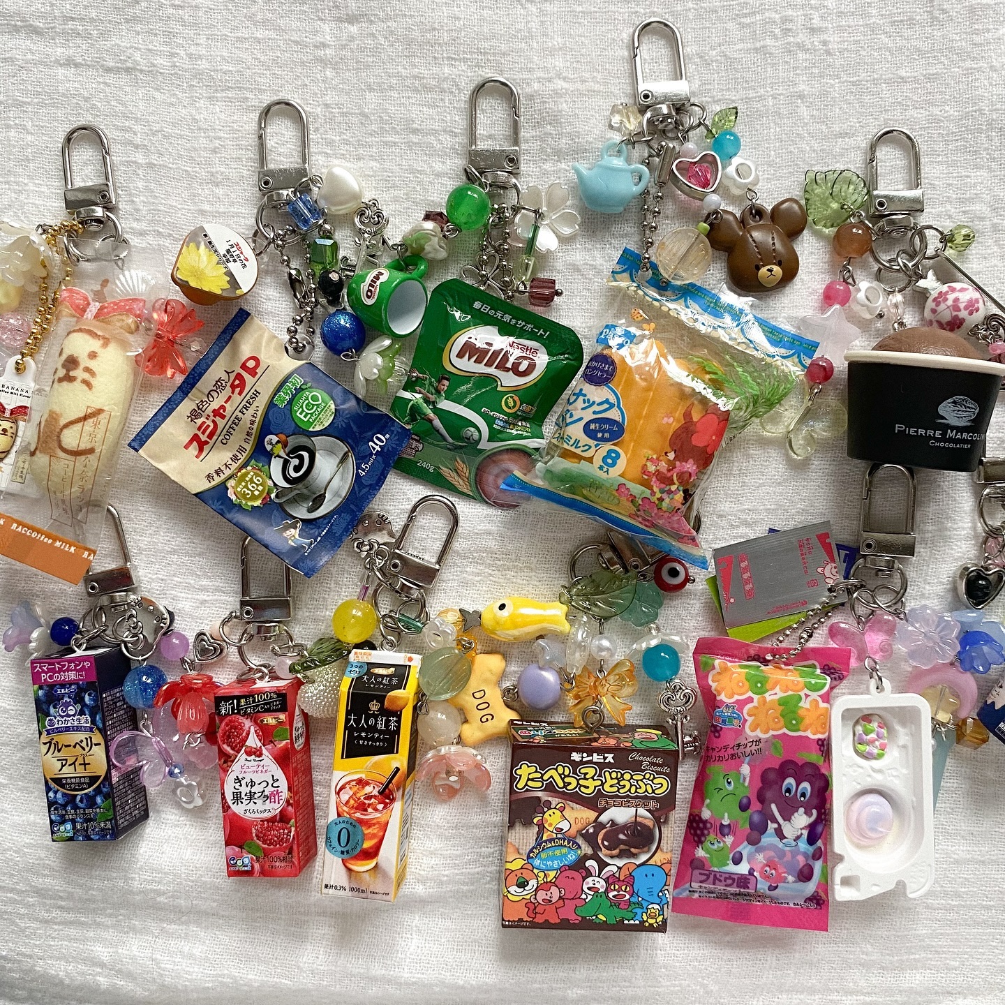 the image displays a collection of keychains adorned with miniature food and snack charms, alongside various beads and embellishments. there are charms representing japanese snacks, milo products, coffee, fruit drinks, animal crackers, grape flavored candies, and a chocolate dessert. other charms include a teapot and a teddy bear.