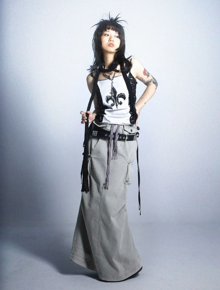 a fullbody shot of a young woman posing against a light grey background. she's wearing a stylish outfit that includes a long, flowing, light grey cargo skirt, a white tank top with a black fleurdelis design, and a black, beaded, layered top.  she has black hair styled in a slightly messy, choppy bob, dark eye makeup, and visible tattoos on her arm.  she is wearing multiple accessories, including a black choker necklace and a studded belt. the overall style is grunge or alternative. the image has a slightly moody, fashion editorial feel.