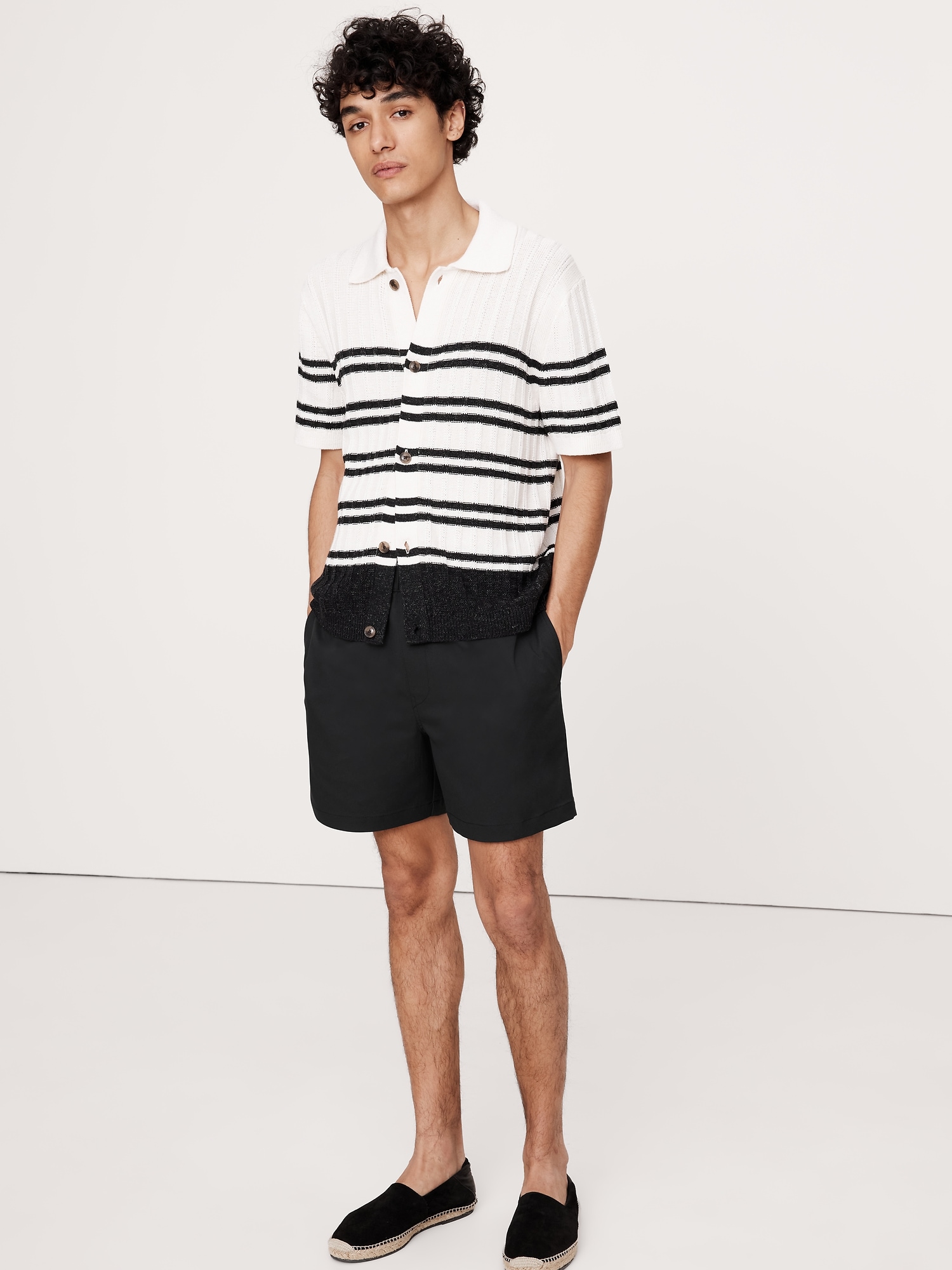 a fulllength studio shot of a man in a white and black striped polo shirt, black shorts, and black espadrilles. he has dark curly hair and stands in front of a white background.