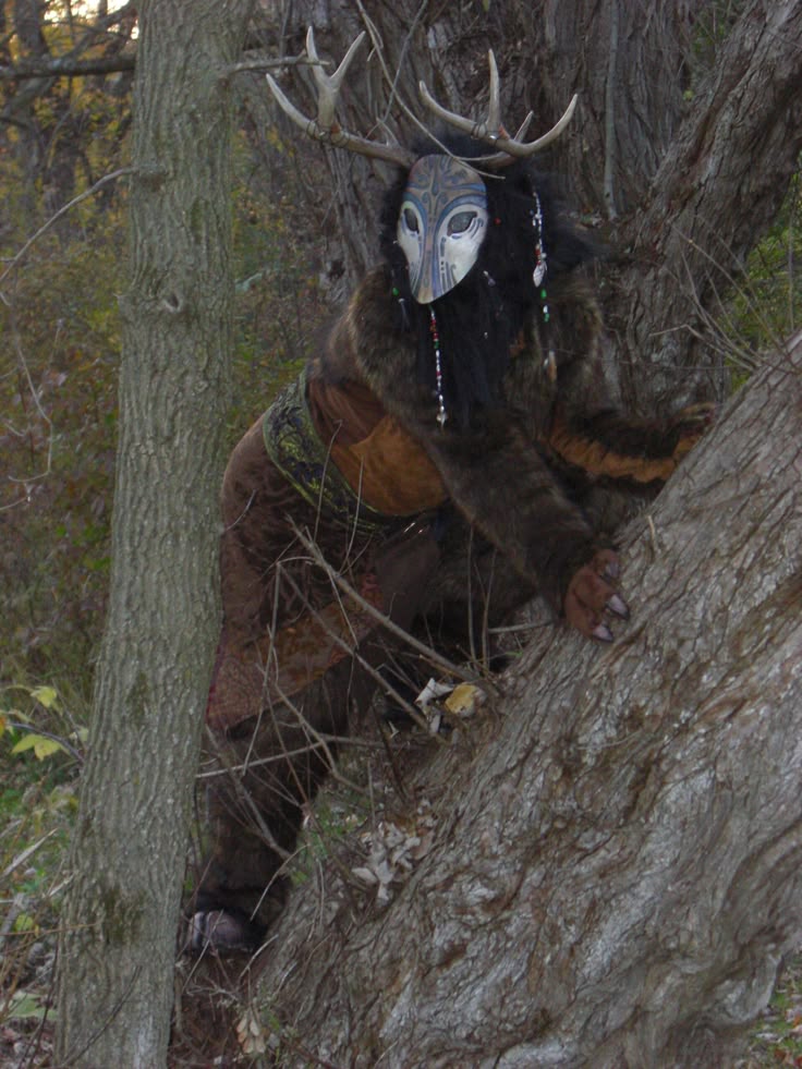 a person wearing a complex costume including a mask, antlers, and furry body suit climbs a large tree in a wooded area. the costume creates the impression of a mythical forest creature. the mask is white and blue with intricate designs, and the antlers add to the creature's mystical appearance. the person is surrounded by trees and leaves, blending into the natural environment.