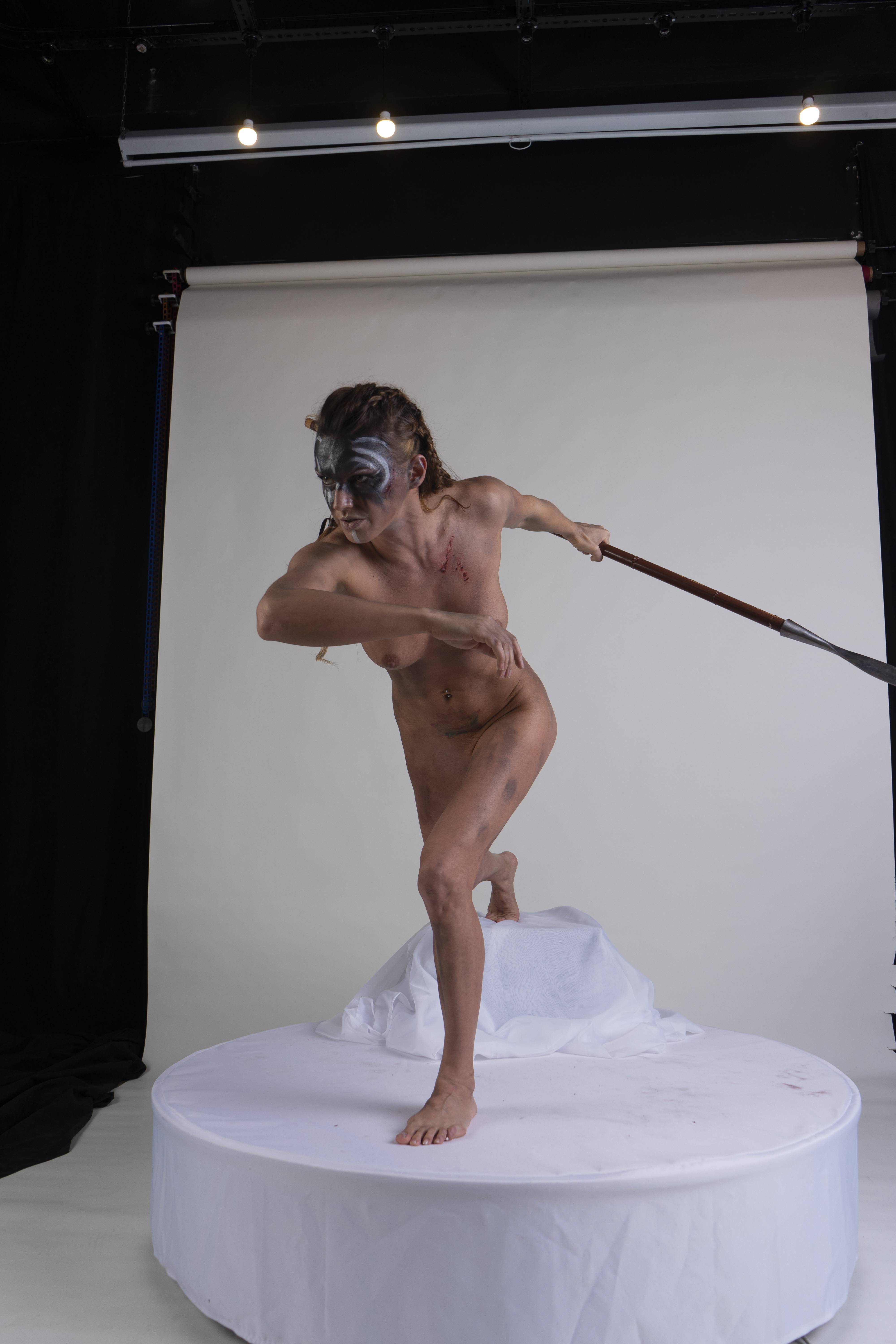 a nude woman with face paint and braided hair stands on a white platform in a studio setting, wielding a spear. she is posed in a dynamic action stance, her expression suggesting determination and aggression. the background features a white backdrop and lighting equipment.