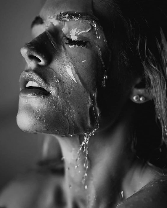 the image is a closeup, black and white portrait of a woman's face and upper body. water is dripping down her face and neck, creating a dramatic and sensual effect.  the lighting is dramatic, highlighting the texture of her skin and the glistening water. her eyes are closed, and her expression is serene and slightly suggestive. the overall mood is elegant and artistic.