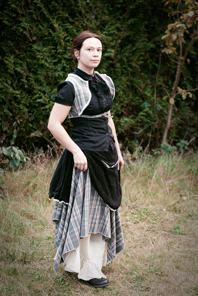 a young woman stands in a grassy area, wearing a stylish outfit. her dress is a combination of black and a beigegrey plaid. she also wears a black waistcoat with white lace detailing. the skirt of the dress is layered, and the overall style is reminiscent of historical or vintage fashion with a touch of modern flair.  the background consists of lush greenery, trees and bushes.  the image has a slightly muted, almost vintage, color tone.