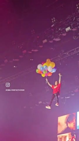 a person is hanging from a bunch of balloons above a concert stage, looking out at the crowd. the person is holding on to the balloons and is suspended in the air. the balloons are a variety of colors, and there are lights behind the person, creating a blurred background. the image was likely taken from a concert, where the person is likely performing for the crowd.