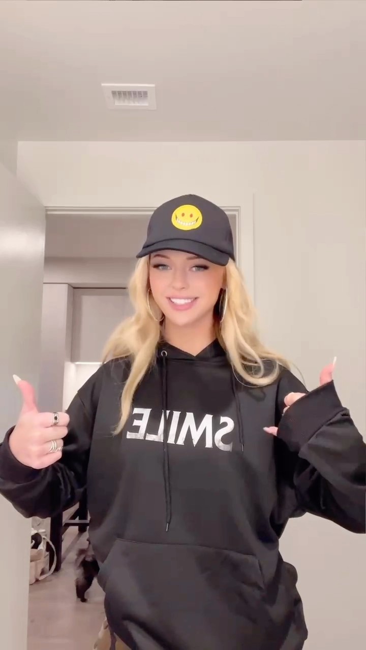 a woman with blonde hair is standing indoors, wearing a black hoodie with the word "smile" printed on it in reverse. she is also wearing a black hat with a smiley face on it. she is giving a thumbs up with both hands. the background shows a doorway and part of a room.
