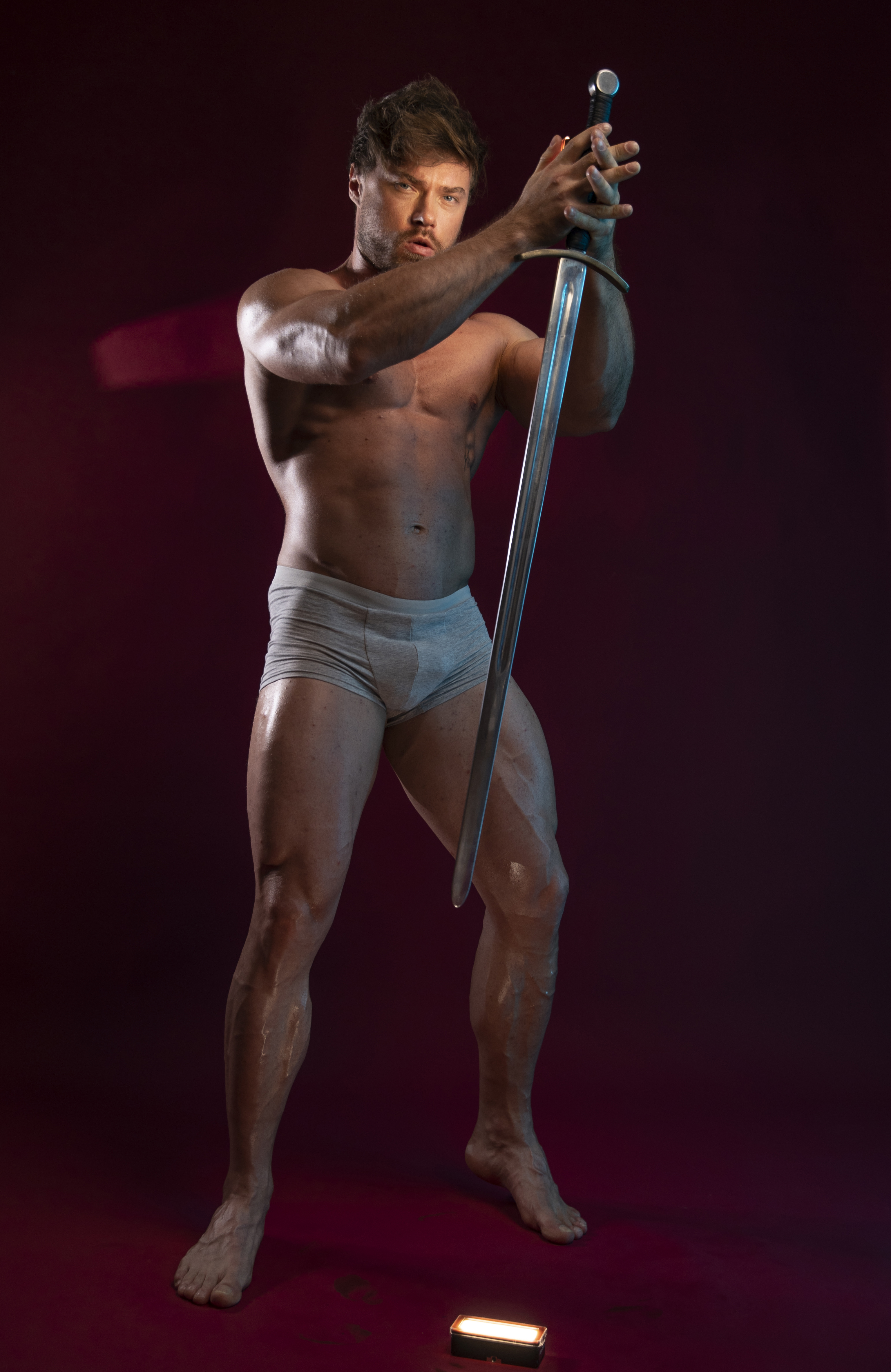 a shirtless, muscular man is holding a sword in a portrait indoors against a red background. he is wearing underwear and standing with the sword in front of him. lighting emphasizes his physique.