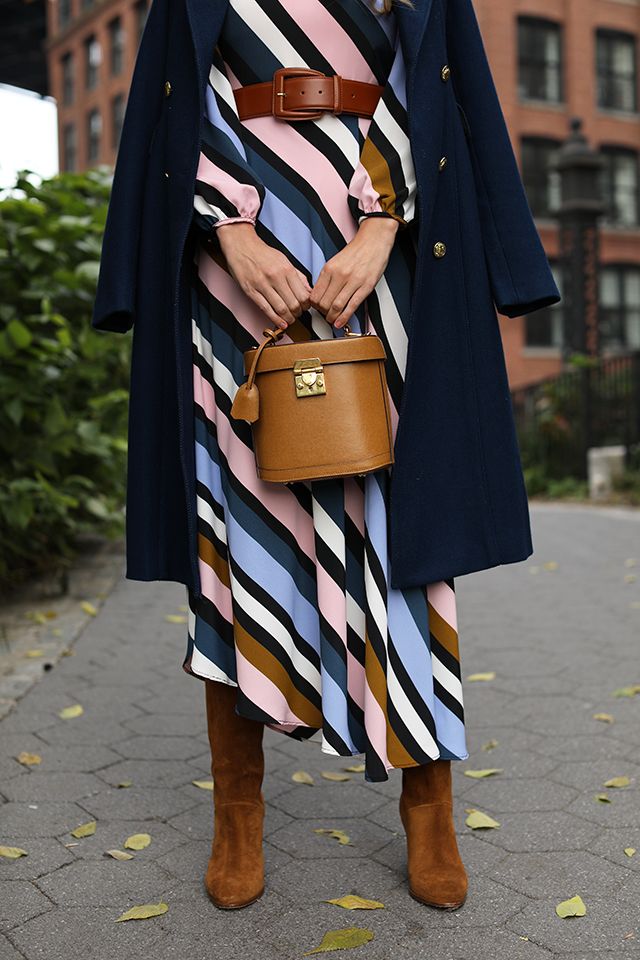 the image shows a stylish woman wearing a multicolored striped midi dress with long sleeves. she's accessorized with a navy blue doublebreasted coat, brown kneehigh suede boots, and a brown leather belt. she carries a small, brown boxshaped handbag. the backdrop is a city street with a brick building visible. the overall aesthetic is fall or autumnal fashion. the photo seems to be taken outdoors during the daytime.