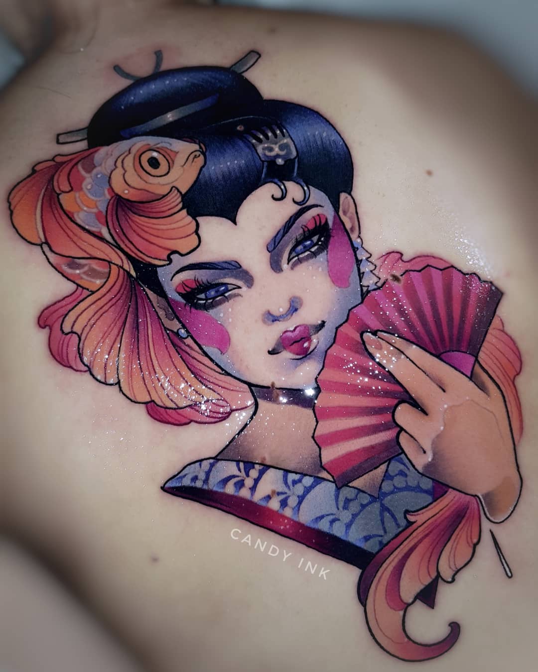 a tattoo of a japanese geisha holding a fan with a goldfish behind her.  the tattoo has a beautiful blend of colors with details in pink, orange, blue, and purple.