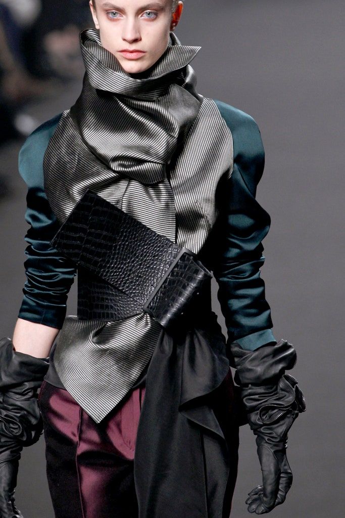 a female model poses on a runway in a chic outfit. she is wearing a black and grey striped scarf that wraps around her neck and is tied in a knot at the front, forming a large bow. over the scarf she wears a teal jacket with large puffy sleeves. the jacket has a wide black belt with a crocodileembossed leather design. the belt cinches the jacket at the waist, adding definition to the outfit. the model is also wearing long black gloves that are adorned with ruffles. the gloves add a touch of drama and elegance to the outfit. the model's hair is pulled back, allowing the focus to be on the statementmaking outfit. the overall effect is a dramatic and stylish look.