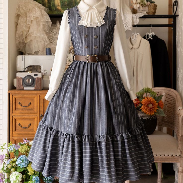 a grey and white striped lolita fashion dress is shown on a mannequin. the dress has a ruffled skirt and a white blouse underneath. the mannequin is wearing a brown belt and a white bow tie. the background is a room with a wooden floor and a wooden cabinet. there are flowers on the floor in front of the mannequin.