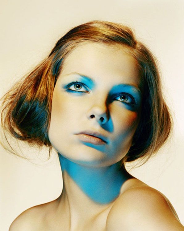 a woman with a striking blue eyeshadow look stares intensely at the camera. she has her hair swept off of her face, and her shoulders are slightly visible. the image is lit from the left side, casting a blue shadow across her face and neck.
