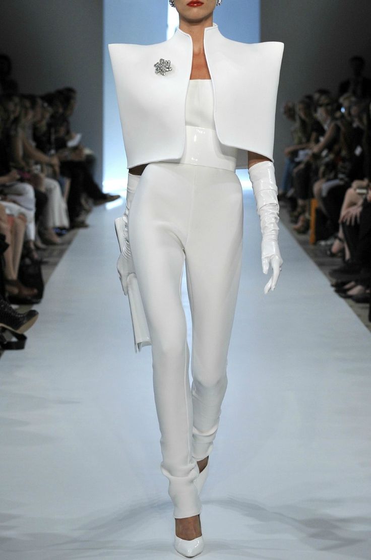 highfashion model strides down a runway in a striking white ensemble.  the outfit comprises a structured, geometric white jacket with exaggerated shoulder pads, a formfitting white jumpsuit, and long white gloves.  a delicate brooch adorns the jacket.  the model's makeup features a bold red lip. the background shows a blurred audience seated on either side of the runway. the overall aesthetic is clean, minimalist, and modern, emphasizing strong architectural lines and a monochromatic color palette.