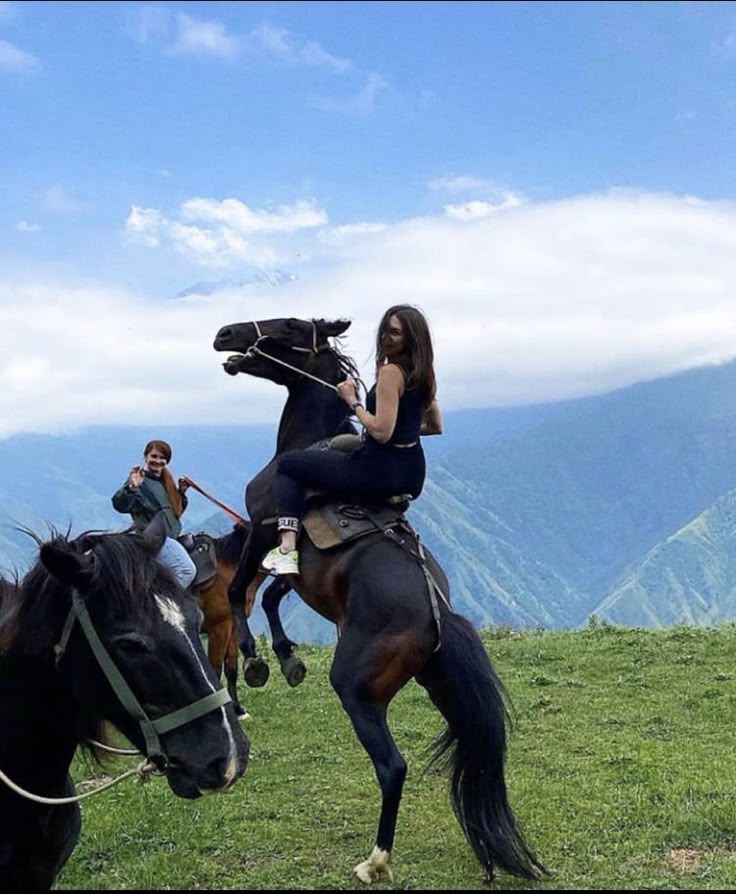the image features two women horseback riding in a mountainous landscape. in the foreground, a woman is riding a black horse that is rearing up on its hind legs. she is dressed in black clothing and white sneakers, and looks back towards the camera. another woman is riding a brown horse in the background. the landscape consists of grassy hills, valleys, and mountains, with a blue sky and clouds overhead. there is also a black horse in the foreground with a white marking on its face.