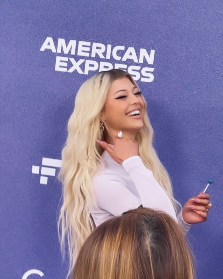 a photo of loren gray at an american express event. she has long blonde hair, is wearing a longsleeve white shirt, and is holding a lollipop. she is smiling and has an earring and a ring on her finger. in the foreground, there is a crowd of people and in the background, there is the american express logo.