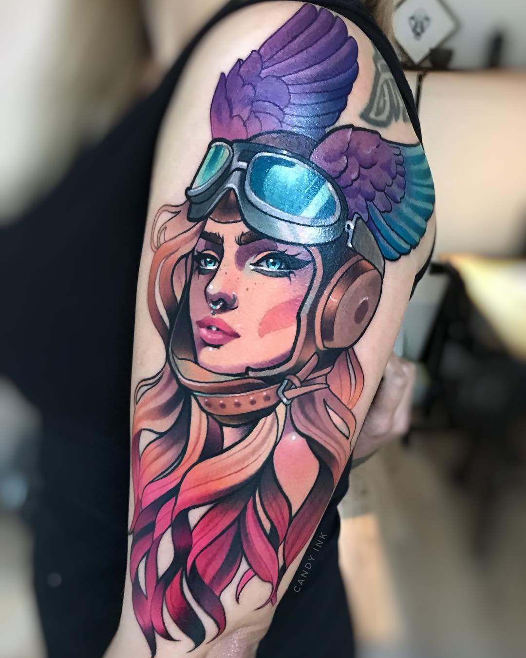the image shows a colorful, detailed tattoo of a woman's portrait on someone's upper arm. the woman in the tattoo has flowing pink and orange hair and is wearing aviator goggles and a helmet with wings attached. the tattoo is done in a realistic style with bold lines and shading.