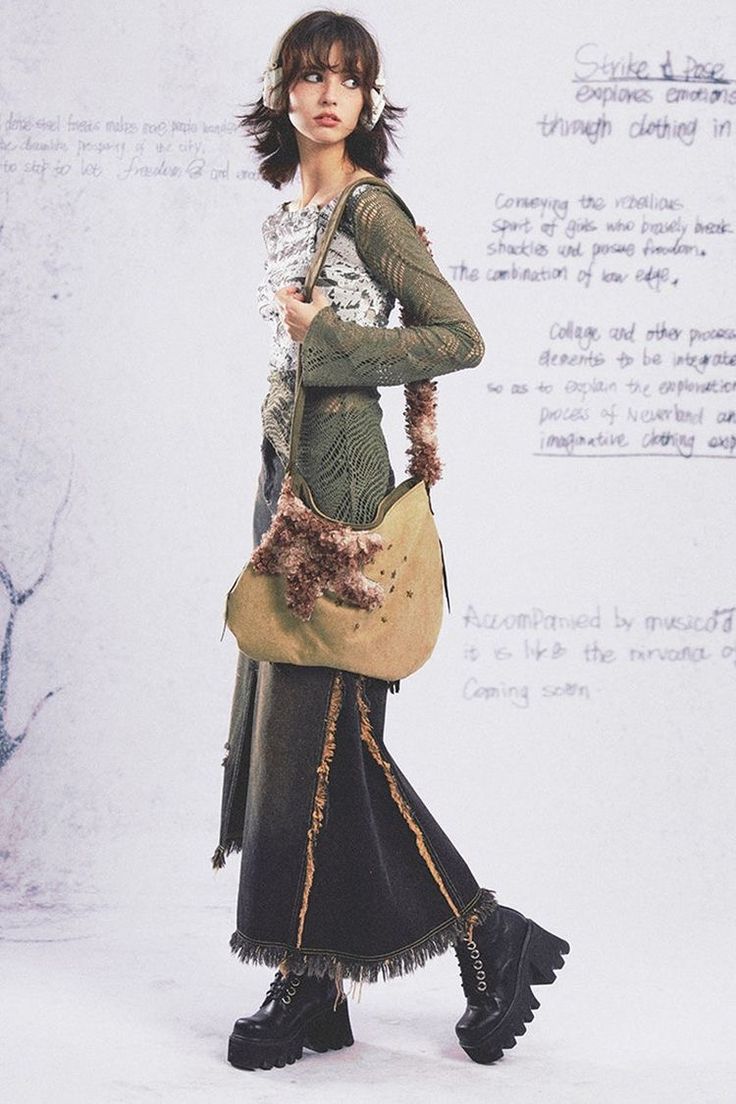 a fashion photograph of a young woman wearing a layered outfit in grunge style. she is wearing a green crocheted top, a long black denim skirt with frayed edges and tan accents, chunky black platform boots, and a large tan hobo bag with fluffy embellishments.  the model has short brown hair and is wearing headphones. the background includes handwritten notes that appear to be design sketches or inspiration notes related to the clothing line or photoshoot. the overall style evokes a 90s grunge aesthetic.