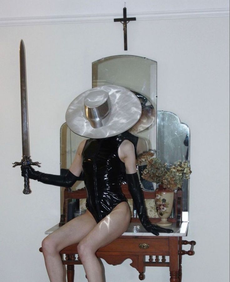 a woman in a black leather outfit, wearing a large silver and white hat, is holding a sword and sitting on a vanity with a mirror in the background. there is a cross hanging above the mirror. she is posing for a picture, and her reflection can be seen in the mirror.