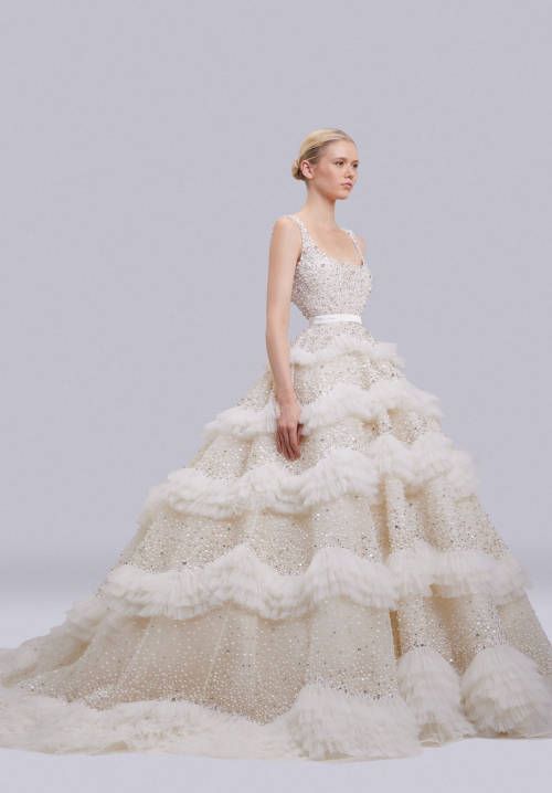 a beautiful bride stands in a white wedding gown. the gown is made of lace and is covered in sparkling embroidery. the bride is wearing her hair up and has a natural look. she looks elegant and glamorous.  the gown has several layers of ruffles.