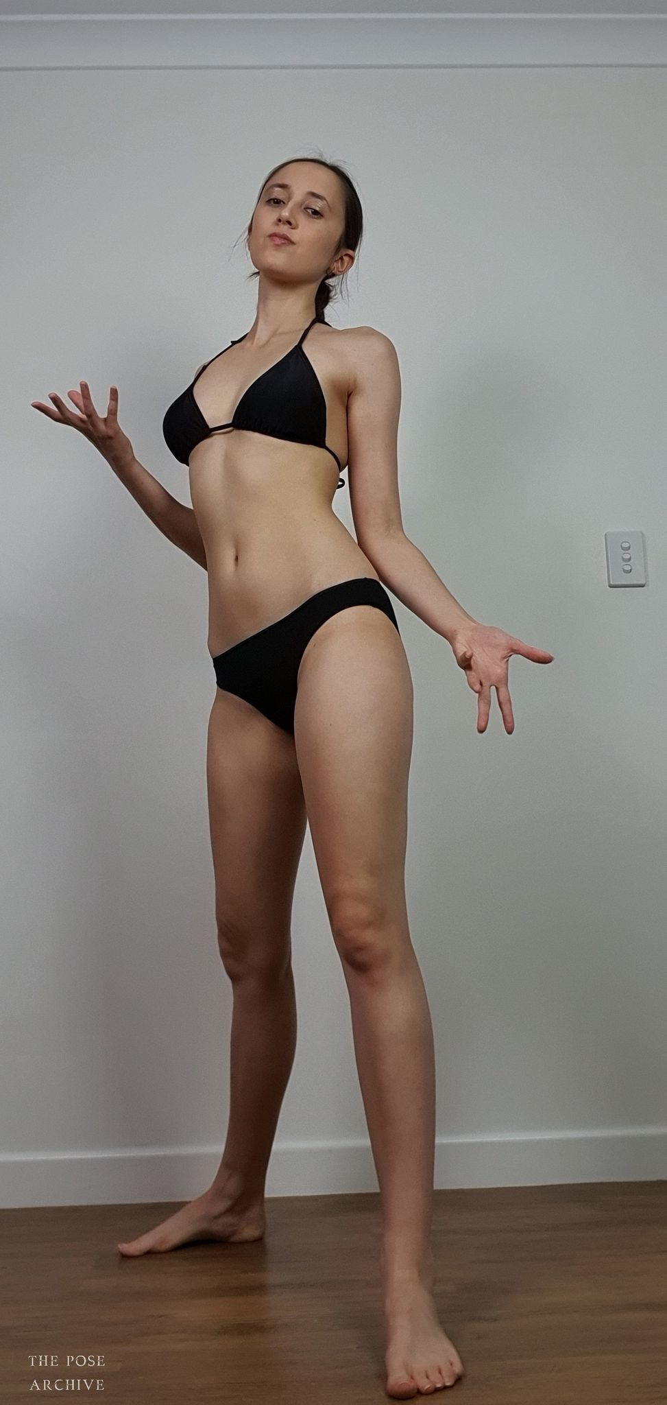 a young woman with a slender build stands barefoot on a hardwood floor against a plain white wall. she's wearing a simple black bikini and is striking a pose with her arms outstretched and palms open, her head tilted slightly upward. the lighting appears to be artificial, casting subtle shadows across her body. the overall aesthetic suggests this image is intended as a pose reference for artists.
