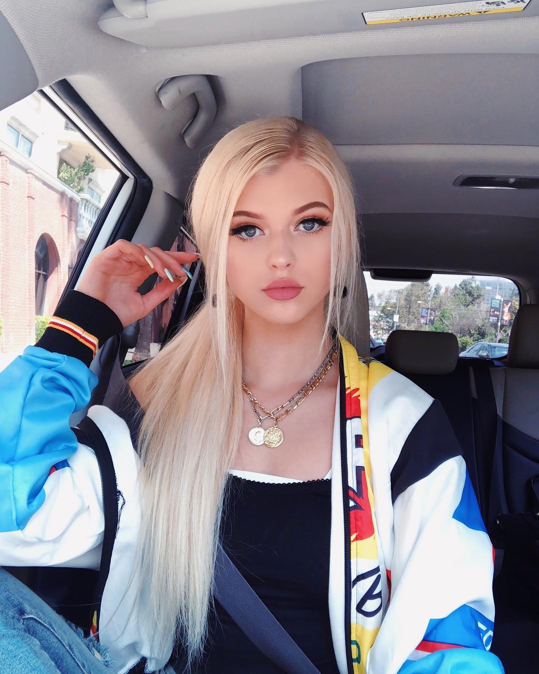 a young woman with blonde hair and blue eyes is sitting in the driver's seat of a car. she's wearing a black tank top, a colorful jacket, and a seatbelt. she's also wearing layered necklaces and has fair skin. the car interior is visible, including the window, auto part, and back seat. the woman is looking at the camera with a neutral expression. she has pink lipstick on. she is loren gray.