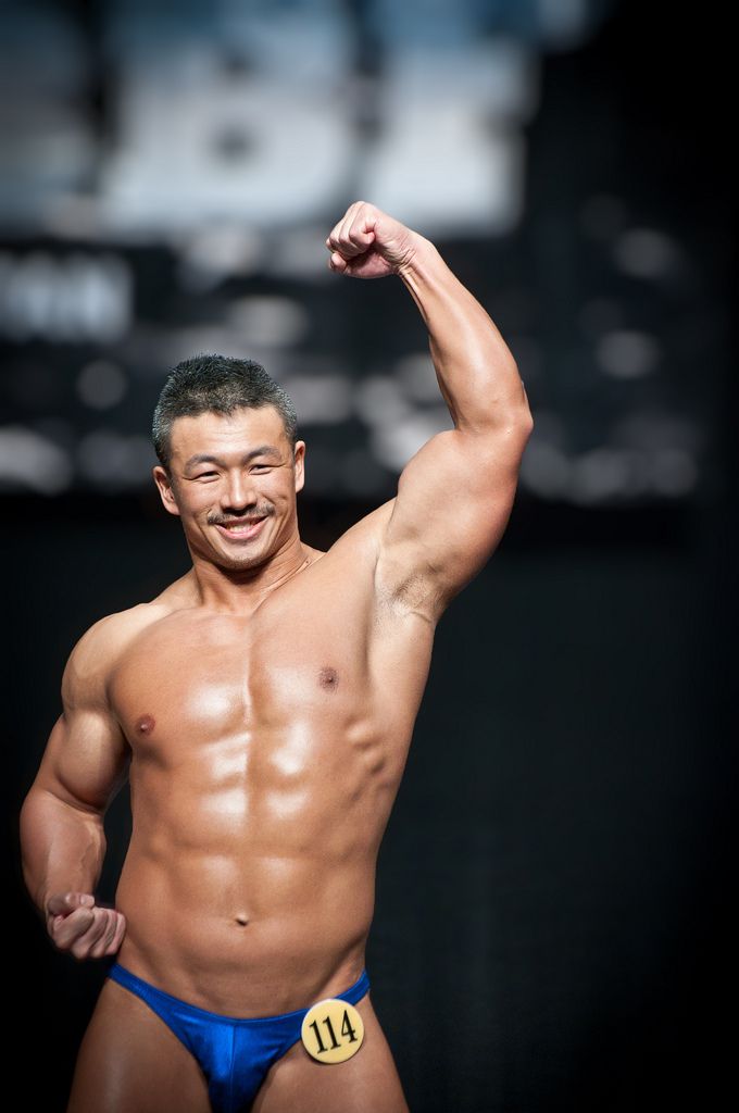 a muscular bodybuilder poses on stage, showcasing his physique in blue swim trunks. he has a competition number, 114, attached to his left hip. the background is blurred, focusing on the subject.