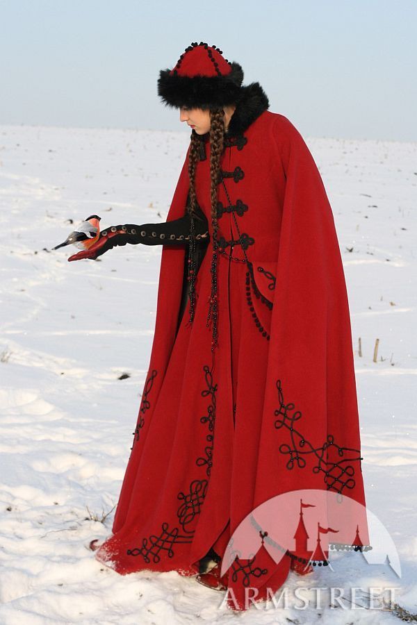 a woman in a long red coat and headdress stands in a snowy landscape, a bird perched on her gloved hand. the coat is adorned with black embroidery, and the headdress is trimmed with fur. the woman has long braids. the bird appears to be a bullfinch. a logo for armstreet is visible in the bottom right corner.
