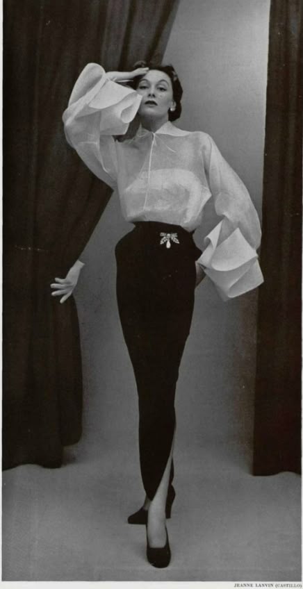 a black and white portrait of a woman in a fashionable pose, showcasing a vintage highfashion outfit. she is wearing a sheer, longsleeved blouse with voluminous, ruffled cuffs, paired with a dark, highwaisted skirt that features a decorative brooch at the waistband and a slit at the front. her hair is styled up, and she strikes an elegant pose with one hand on her head and the other extended. the photograph is likely from a vintage fashion magazine or catalog, credited to jeanne lanvin.